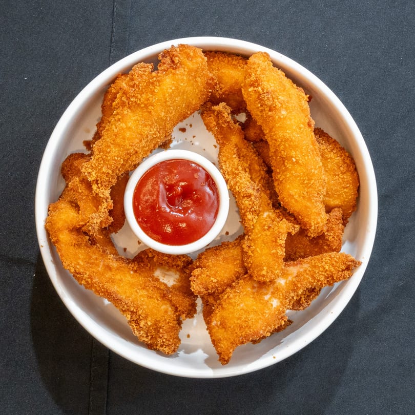 Crispy golden-brown chicken tenders with a side of bright red ketchup in a white bowl.