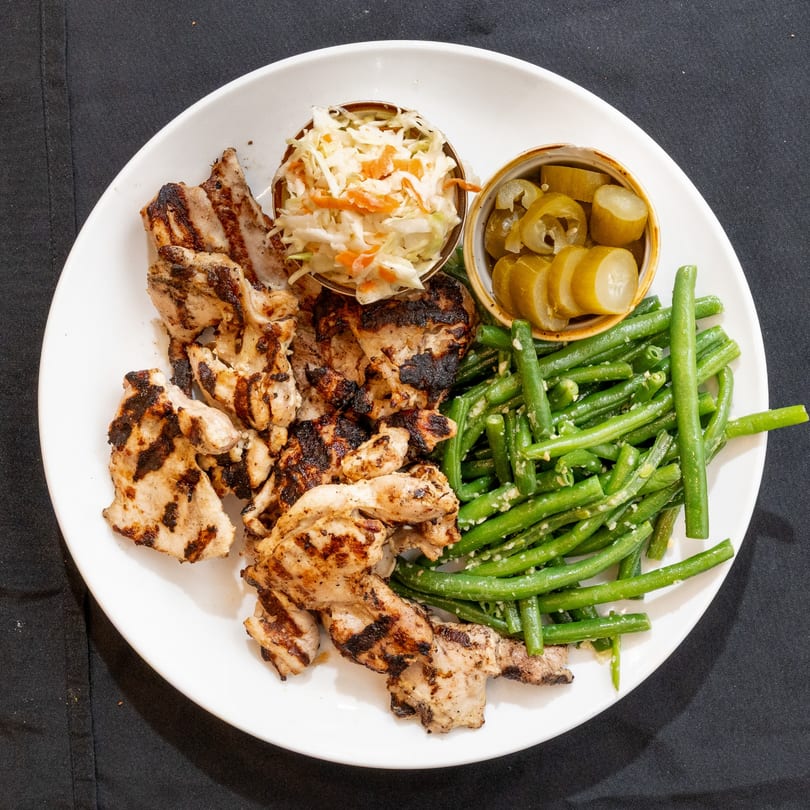 Grilled chicken pieces, green beans, coleslaw, and sliced pickles on a white plate.