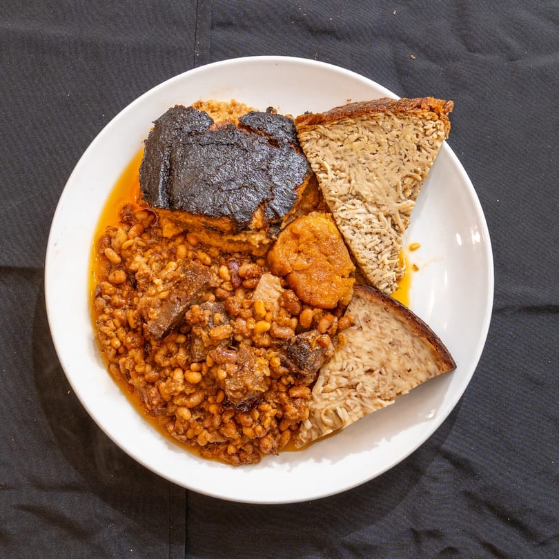 Hearty kosher cholent with beans, meat, kugel, and a dark crust on a white plate.