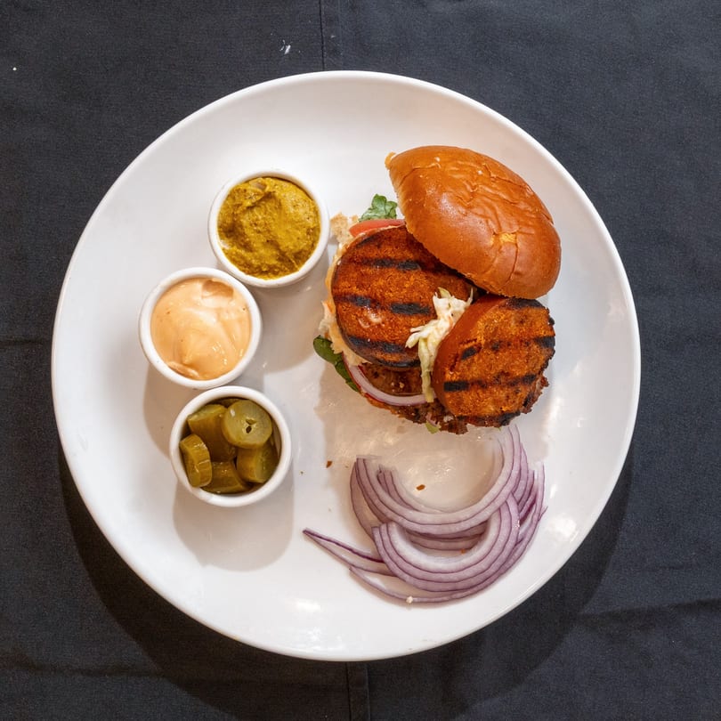 Grilled kosher burger with red onion, pickles, mustard, and sauce on a white plate.