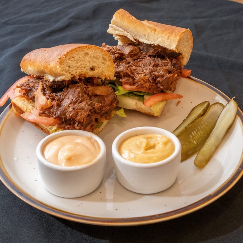 Two Zilus Essen kosher pulled beef sandwiches with lettuce, tomato, pickles, and two dipping sauces.