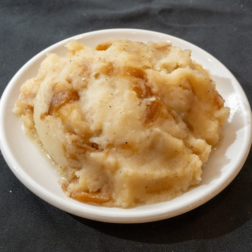 A white bowl filled with creamy mashed potatoes, mixed with caramelized onions and a savory sauce.