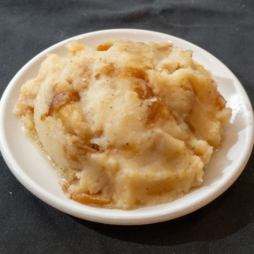 A white bowl filled with creamy mashed potatoes, mixed with caramelized onions and a savory sauce.